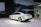 A sleek, white sedan is displayed on a showroom floor with a large digital backdrop featuring a close-up of the car's headlight design. The environment appears modern and minimalistic, and a QR code is visible on the left side of the image. Two people are casually observing the scene from a distance.