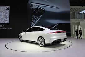 A sleek, white sedan is displayed on a showroom floor with a large digital backdrop featuring a close-up of the car's headlight design. The environment appears modern and minimalistic, and a QR code is visible on the left side of the image. Two people are casually observing the scene from a distance.