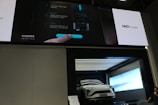 A white electric vehicle is showcased inside a modern, illuminated display booth. Above the vehicle is a large screen displaying information in both Chinese and English, along with the branding 'NIO Power.' The screen shows interface instructions involving a hand interacting with a digital display. To the side, a person is partially visible.