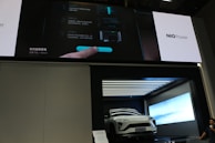 A white electric vehicle is showcased inside a modern, illuminated display booth. Above the vehicle is a large screen displaying information in both Chinese and English, along with the branding 'NIO Power.' The screen shows interface instructions involving a hand interacting with a digital display. To the side, a person is partially visible.