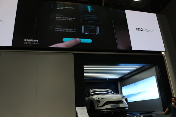 A white electric vehicle is showcased inside a modern, illuminated display booth. Above the vehicle is a large screen displaying information in both Chinese and English, along with the branding 'NIO Power.' The screen shows interface instructions involving a hand interacting with a digital display. To the side, a person is partially visible.
