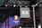 Hanging signage with bold, stylized lettering is displayed in a large indoor venue. Below, a digital display showcases a logo featuring a lion's head and text with vibrant blue and red lighting effects. The ceiling is lined with intricate lighting fixtures and metal trusses.
