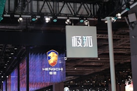 Hanging signage with bold, stylized lettering is displayed in a large indoor venue. Below, a digital display showcases a logo featuring a lion's head and text with vibrant blue and red lighting effects. The ceiling is lined with intricate lighting fixtures and metal trusses.