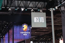 Hanging signage with bold, stylized lettering is displayed in a large indoor venue. Below, a digital display showcases a logo featuring a lion's head and text with vibrant blue and red lighting effects. The ceiling is lined with intricate lighting fixtures and metal trusses.