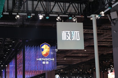 Hanging signage with bold, stylized lettering is displayed in a large indoor venue. Below, a digital display showcases a logo featuring a lion's head and text with vibrant blue and red lighting effects. The ceiling is lined with intricate lighting fixtures and metal trusses.