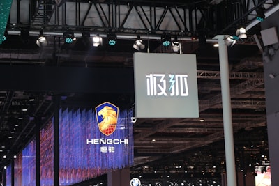 Hanging signage with bold, stylized lettering is displayed in a large indoor venue. Below, a digital display showcases a logo featuring a lion's head and text with vibrant blue and red lighting effects. The ceiling is lined with intricate lighting fixtures and metal trusses.