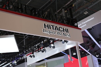 Industrial expo booth featuring a prominent sign with 'Hitachi' and the slogan 'Inspire the Next.' The design is modern and sleek, incorporating various lighting fixtures and a red, black, and white color scheme. The structure appears to be part of a larger exhibition with multiple technology displays and installations in the background.