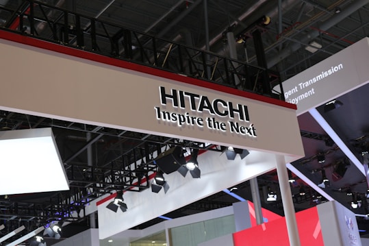 Industrial expo booth featuring a prominent sign with 'Hitachi' and the slogan 'Inspire the Next.' The design is modern and sleek, incorporating various lighting fixtures and a red, black, and white color scheme. The structure appears to be part of a larger exhibition with multiple technology displays and installations in the background.
