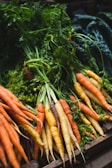 Multiple bunches of freshly picked carrots, in various colors including orange, yellow, and purple, with their green tops still attached. The carrots are laid out together in a wooden box or crate with some leafy greens visible in the background.