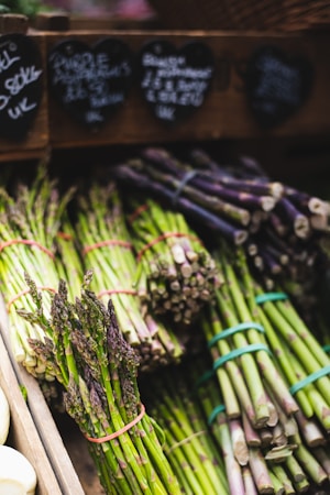 Bundles of fresh green and purple asparagus are displayed on a market stand. Each bundle is tied with a rubber band. Behind the asparagus are chalkboard price tags in the shape of hearts.