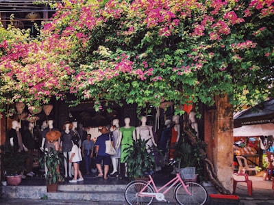 A vibrant street scene with colorful pink Bougainvillea flowers cascading over a shop entrance, where mannequins dressed in various outfits are displayed. People are casually walking up the steps into the shop, and a pink bicycle with a basket is parked nearby surrounded by potted plants.