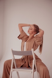 A person with long hair sits on a chair, wearing a loose, beige outfit. The setting is minimal, with a neutral-toned backdrop, giving a soft and serene feeling. The person has their eyes closed, with their hands raised, likely running fingers through their hair.