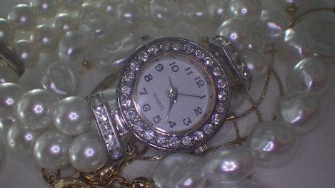 A jeweled watch with a round face surrounded by sparkling rhinestones is displayed amidst a pile of shiny, white pearls. The numbers on the dial are bold and clearly printed, and the watch hands point to a specific time. The lustrous pearls are strung together, creating a luxurious and elegant composition.