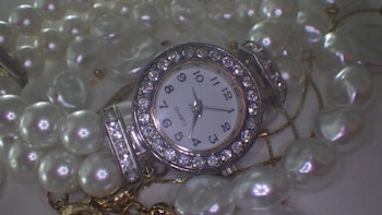 A jeweled watch with a round face surrounded by sparkling rhinestones is displayed amidst a pile of shiny, white pearls. The numbers on the dial are bold and clearly printed, and the watch hands point to a specific time. The lustrous pearls are strung together, creating a luxurious and elegant composition.