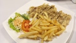 A white plate containing crispy fried chicken strips, a serving of golden French fries, and a side salad with shredded cabbage, lettuce, sliced cucumber, and a tomato slice topped with a dollop of creamy dressing.