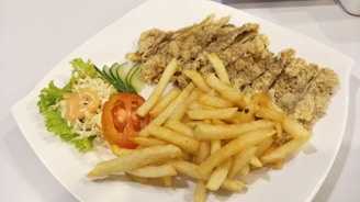 A white plate containing crispy fried chicken strips, a serving of golden French fries, and a side salad with shredded cabbage, lettuce, sliced cucumber, and a tomato slice topped with a dollop of creamy dressing.