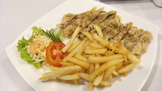 A white plate containing crispy fried chicken strips, a serving of golden French fries, and a side salad with shredded cabbage, lettuce, sliced cucumber, and a tomato slice topped with a dollop of creamy dressing.