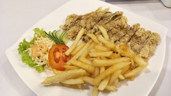 A white plate containing crispy fried chicken strips, a serving of golden French fries, and a side salad with shredded cabbage, lettuce, sliced cucumber, and a tomato slice topped with a dollop of creamy dressing.