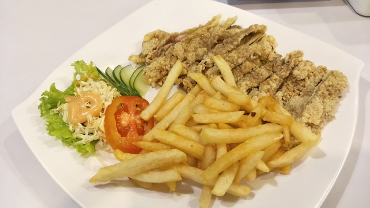 A white plate containing crispy fried chicken strips, a serving of golden French fries, and a side salad with shredded cabbage, lettuce, sliced cucumber, and a tomato slice topped with a dollop of creamy dressing.