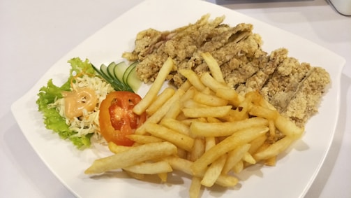 A white plate containing crispy fried chicken strips, a serving of golden French fries, and a side salad with shredded cabbage, lettuce, sliced cucumber, and a tomato slice topped with a dollop of creamy dressing.