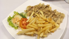 A white plate containing crispy fried chicken strips, a serving of golden French fries, and a side salad with shredded cabbage, lettuce, sliced cucumber, and a tomato slice topped with a dollop of creamy dressing.
