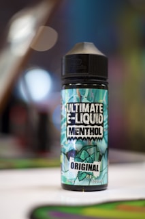 A bottle of e-liquid is prominently displayed, featuring a label with the text 'Ultimate E-Liquid Menthol Original.' The label is decorated with images of mint leaves and an icy background, suggesting a cooling menthol flavor. The bottle cap is black, and the background is blurry, with bokeh lights, creating a focused and isolated view of the product.