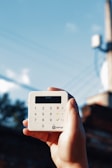 A hand is holding a small, white, square-shaped card reader against a backdrop of a clear blue sky with some power lines. The device has a digital display and a keypad with numerical and functional buttons.