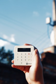 A hand is holding a small, white, square-shaped card reader against a backdrop of a clear blue sky with some power lines. The device has a digital display and a keypad with numerical and functional buttons.