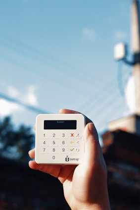 A hand is holding a small, white, square-shaped card reader against a backdrop of a clear blue sky with some power lines. The device has a digital display and a keypad with numerical and functional buttons.