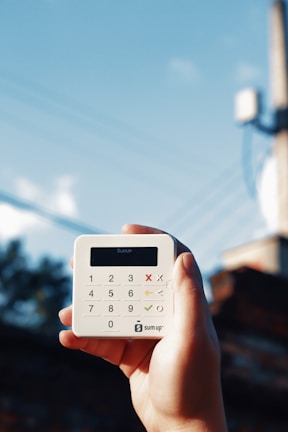 A hand is holding a small, white, square-shaped card reader against a backdrop of a clear blue sky with some power lines. The device has a digital display and a keypad with numerical and functional buttons.