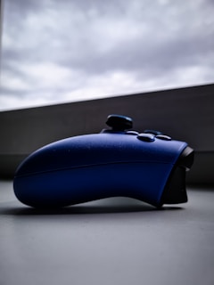 A blue gaming controller placed on a grey surface with a cloudy sky visible in the background. The controller is viewed from a side angle, highlighting its ergonomic shape and buttons.