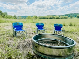 Three blue folding camping chairs are set up on a grassy field with a large metal fire ring or container in the foreground. The background shows a vast open landscape under a bright sky filled with scattered clouds.