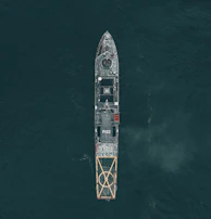 Aerial view of a large naval vessel navigating through dark ocean waters, with detailed features such as a helipad at the stern and the marking P132 visible. The ship's structure stands out against the contrasting dark sea.