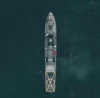 Aerial view of a large naval vessel navigating through dark ocean waters, with detailed features such as a helipad at the stern and the marking P132 visible. The ship's structure stands out against the contrasting dark sea.