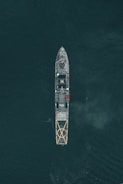 Aerial view of a large naval vessel navigating through dark ocean waters, with detailed features such as a helipad at the stern and the marking P132 visible. The ship's structure stands out against the contrasting dark sea.