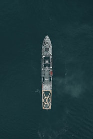 Aerial view of a large naval vessel navigating through dark ocean waters, with detailed features such as a helipad at the stern and the marking P132 visible. The ship's structure stands out against the contrasting dark sea.
