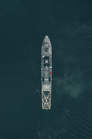 Aerial view of a large naval vessel navigating through dark ocean waters, with detailed features such as a helipad at the stern and the marking P132 visible. The ship's structure stands out against the contrasting dark sea.