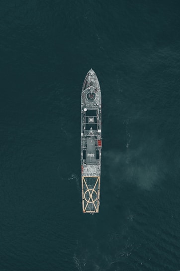 Aerial view of a large naval vessel navigating through dark ocean waters, with detailed features such as a helipad at the stern and the marking P132 visible. The ship's structure stands out against the contrasting dark sea.