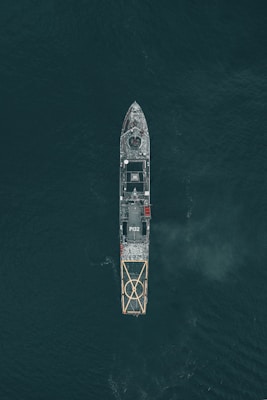 Aerial view of a large naval vessel navigating through dark ocean waters, with detailed features such as a helipad at the stern and the marking P132 visible. The ship's structure stands out against the contrasting dark sea.