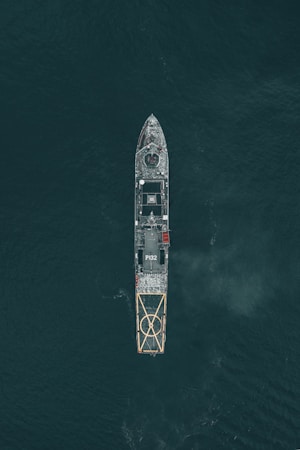 Aerial view of a large naval vessel navigating through dark ocean waters, with detailed features such as a helipad at the stern and the marking P132 visible. The ship's structure stands out against the contrasting dark sea.
