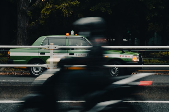 A green taxi is parked on the side of the road, while a person on a motorcycle rides past in the foreground, partially blurred. The scene is set against a backdrop of dark, leafy trees.