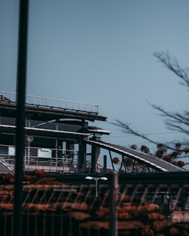 A modern building with steel and glass architecture, featuring rooftop railings and sleek structural lines. In the foreground, blurred fencing and foliage can be seen, with hints of trees and industrial elements.
