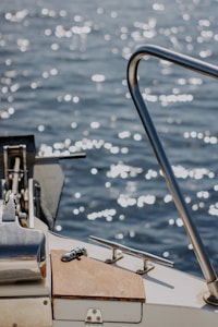 The image captures a close-up view of a boat deck with metallic railings and clear water reflecting sunlight in the background. The surface of the water sparkles with light, creating a serene and reflective atmosphere. The wooden and metallic elements on the boat are sharply in focus, showcasing their texture and design.