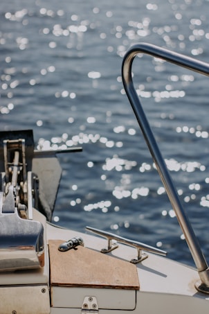 The image captures a close-up view of a boat deck with metallic railings and clear water reflecting sunlight in the background. The surface of the water sparkles with light, creating a serene and reflective atmosphere. The wooden and metallic elements on the boat are sharply in focus, showcasing their texture and design.