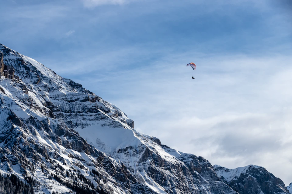 Paraglider soaring high above snow-covered mountain range with clear blue sky in background