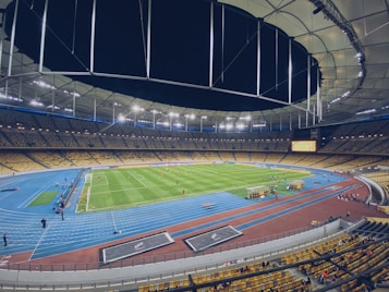 A large, illuminated stadium with a green soccer field and surrounding blue running track. The stands are filled with yellow seats, and a few spectators and players are visible on the field and sidelines. Bright lights hang from the ceiling, casting a well-lit atmosphere.