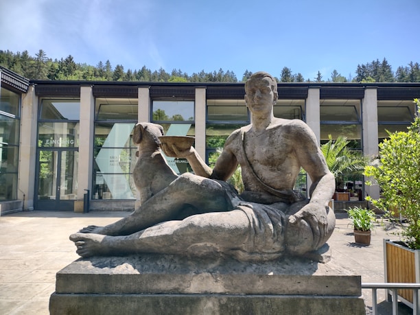 A stone sculpture depicting a partially reclined male figure holding an object, set in an outdoor area with a modern building and glass windows in the background. The scene is surrounded by greenery, including trees and potted plants, under a clear blue sky.