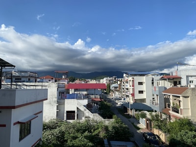 A residential neighborhood featuring modern houses with flat roofs and terraces, located under a sky filled with fluffy white clouds. The scene includes a row of buildings in various shades of white, beige, and red with lush greenery surrounding the area. In the background, distant mountains can be seen under the dramatic cloud-filled sky.