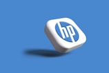 A three-dimensional, stylized version of the HP logo appears on a solid blue background. The logo is displayed in white and blue colors, with a subtle shadow cast beneath it, giving a floating effect.