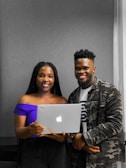 Two people smiling while standing close together, with one holding a laptop displaying an Apple logo. The person on the left is wearing a purple off-shoulder top, and the person on the right is wearing a camouflage jacket over a t-shirt.